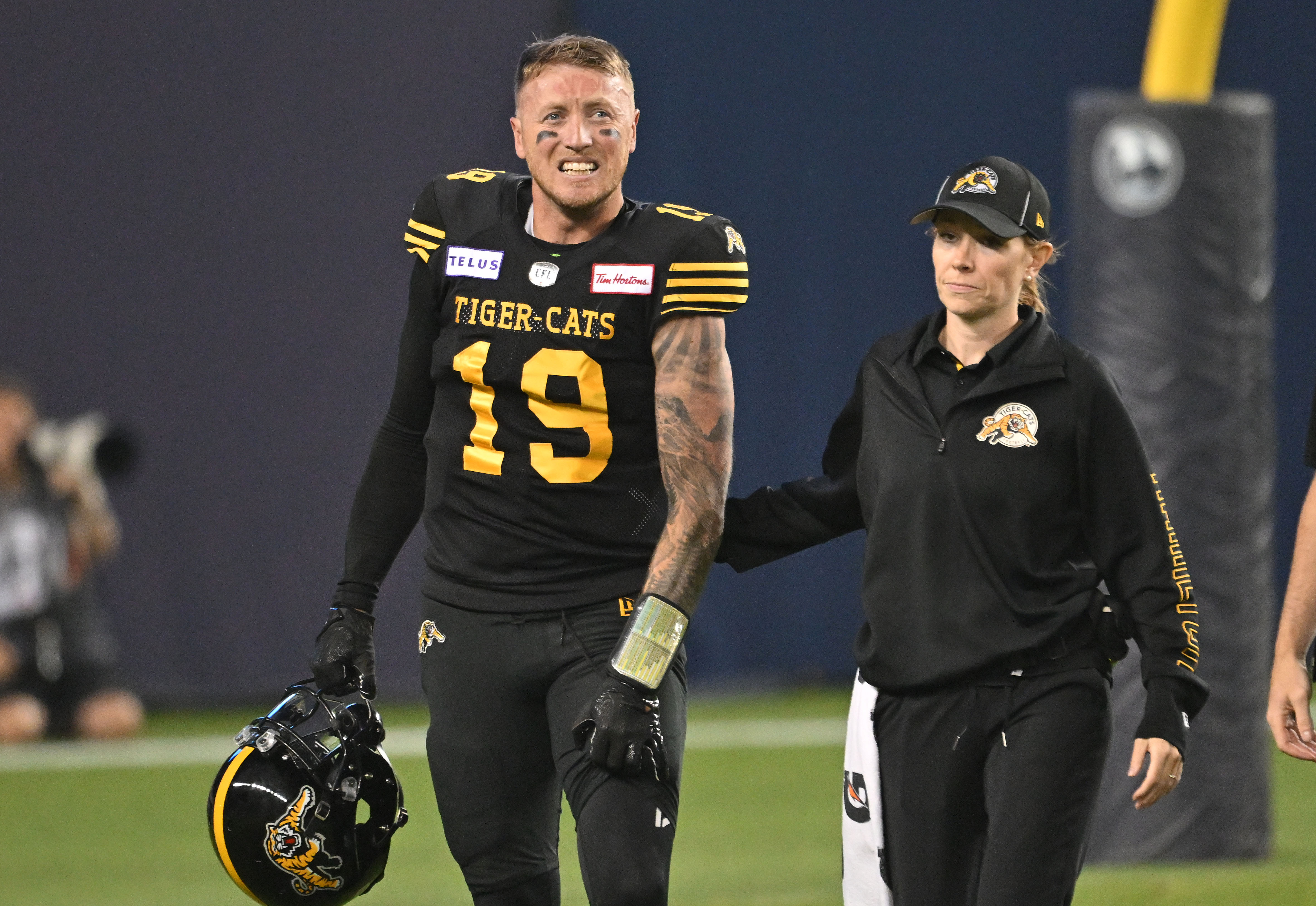 Jun 18, 2023; Toronto, Ontario, CAN; Hamilton Tiger-Cats quarterback Bo Levi Mitchell (19) is escorted off the field by team head therapist Claire Tofflemire after suffering an injury against the Toronto Argonauts in the fourth quarter at BMO Field. Mandatory Credit: Dan Hamilton-USA TODAY Sports